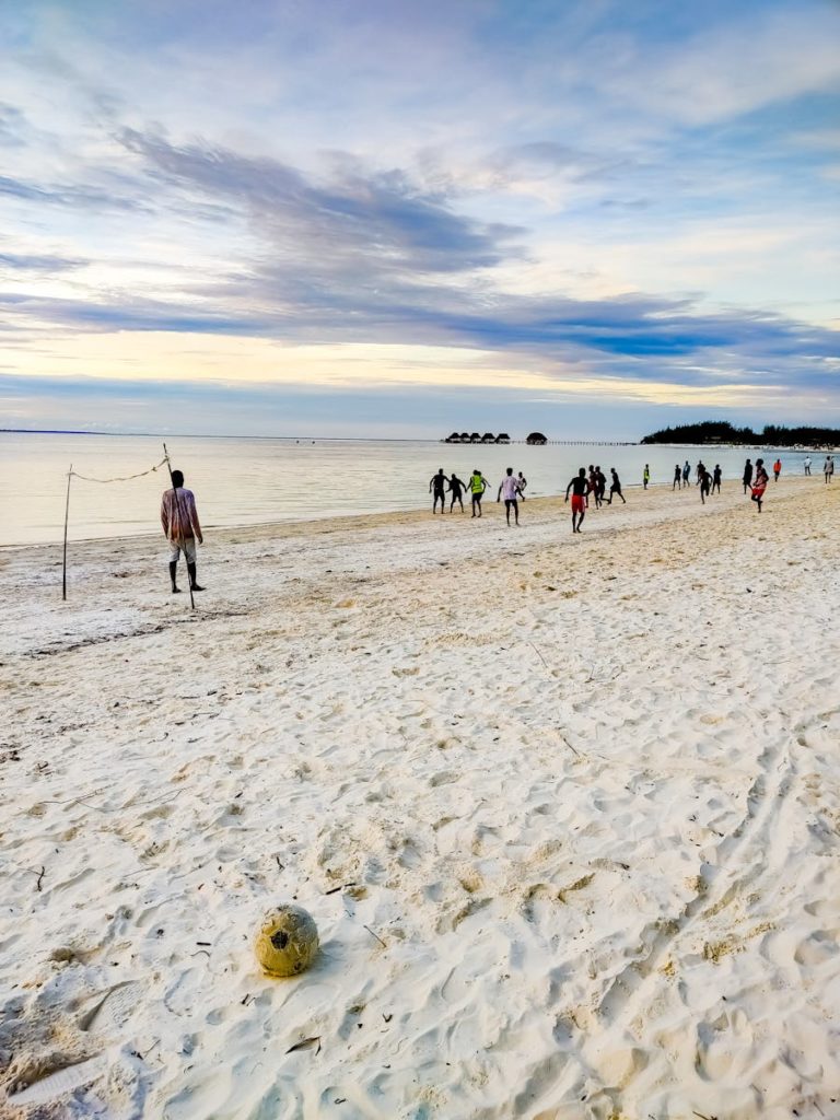 A group of people playing soccer on the sandy shores of Bwejuu, Tanzania during sunset.